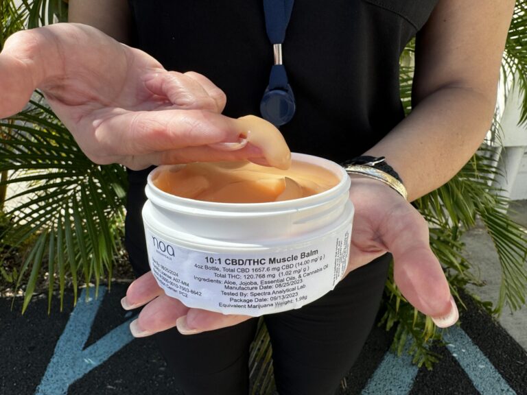 A woman holding a jar of CBD muscle balm in front of a palm tree.