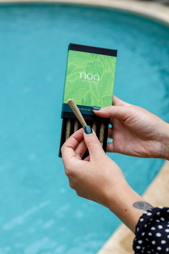 A person holding a pack of noa botanicals pre-rolled cannabis joints by a pool, showcasing a product with a green leafy design.