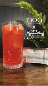 A vibrant red THC infused Fruit Punch Drink Mix in a geometric glass, positioned on a table with a green plant in the background, featuring labels from noa botanicals and hawaiian alchemist.