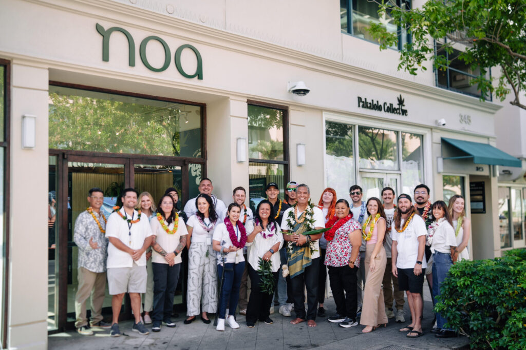 A group of people wearing leis stands in front of Noa Botanicals cannabis dispensary posing for a group photo on a sunny day, celebrating career achievements and community spirit on Oahu Hawaii