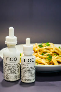 Two bottles of Noa Botanicals Cannabis infused Shoyu and sesame oil are placed in front of a plate of pasta garnished with herbs, perfect inspiration for your next Cannabis edible recipe.