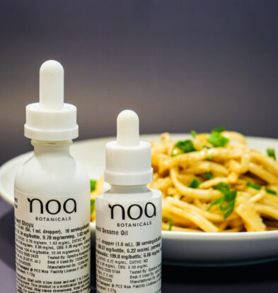 Two bottles of Noa Botanicals Cannabis infused Shoyu and sesame oil are placed in front of a plate of pasta garnished with herbs, perfect inspiration for your next Cannabis edible recipe.